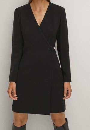 Woman wearing a black long-sleeve wrap dress with a V-neckline and a small buckle detail at the waist, paired with black knee-high boots.