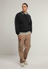 Černý pletený pulovr s bílým límcem, s logem na nášivce; v kombinaci s béžovými cargo kalhotami a bílými teniskami; casual střih.