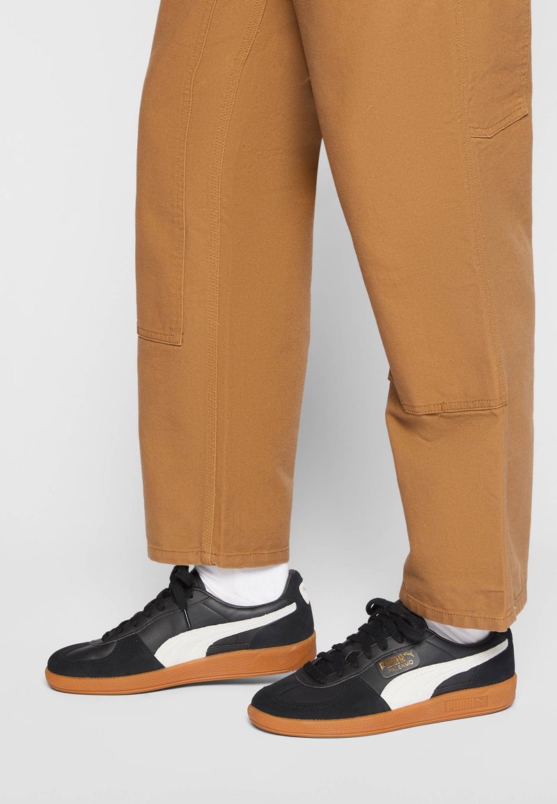 Černobílé tenisky Puma s gumovými podrážkami, nošené s bílými ponožkami a hnědými kalhotami na jednoduchém šedém pozadí.