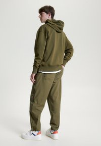 Olivgrüner Hoodie mit einer Kordelzugkapuze, kombiniert mit lockeren olivgrünen Cargo-Hosen. Weiße Sneaker mit roten und blauen Akzenten.