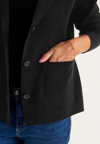Sort tekstureret cardigan med en slå-krave, to knapper og en sidelomme. Kombineret med blå jeans og en sort top.