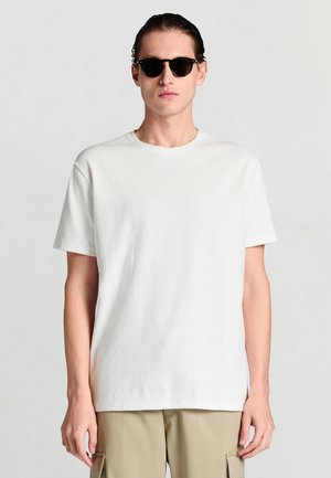 T-shirt bianca testurizzata a maniche corte, collo tondo, abbinata a occhiali da sole neri. Focus su design minimalista e palette di colori neutri.