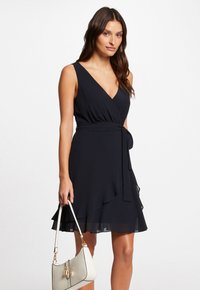 Schwarzes Wickelkleid mit V-Ausschnitt, tailliertem Oberteil, Taillengurt und geschichteter Rüsche am Saum. Das Model hält eine cremefarbene Handtasche mit schwarzen Akzenten.