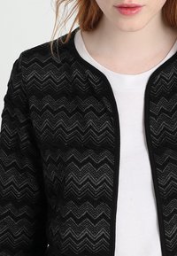 Černá bunda s cikcak vzorem a texturovaným povrchem, doplněná o solidní černé zakončení, nošená přes bílou košili. Hlavní designové prvky zahrnují kulatý výstřih.
