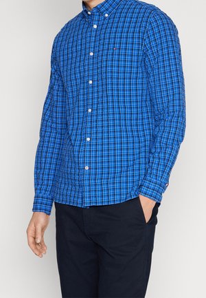 Mann trägt ein blau kariertes, langärmeliges Hemd mit weißen Knöpfen und schwarzen Hosen, eine Hand in der Tasche, vor einem schlichten Hintergrund.