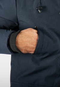 Veste bleu marine en tissu texturé, avec une main dans la poche latérale, et des poignets en tricot côtelé pour plus de confort.