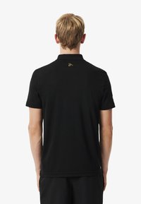 Czarna koszulka polo z krótkim rękawem, prążkowanym kołnierzykiem i małym złotym logo na górnej części pleców. Miękka tkanina, czyste linie i swobodny design.