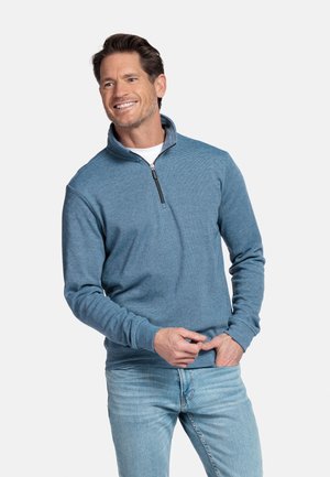 Lachende man die een blauwe pullover met kwartritssluiting en lichtblauwe spijkerbroek draagt, staand met één hand in zijn zak tegen een effen achtergrond.