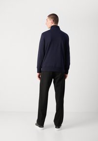 Navy pullover with high collar and ribbed hem, paired with black trousers and dark sneakers, viewed from the back against a white background.