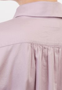Camicia lavanda chiara con colletto classico, caratterizzata da un design con retro plissettato e una texture del tessuto morbida e liscia.