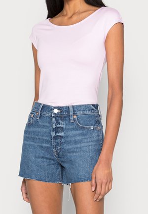 Kobieta nosząca jasnoróżowy top z krótkimi rękawami i wysokimi, niebieskimi, jeansowymi szortami z surowym wykończeniem na jednolitym tle.
