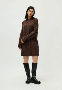 Femeie în picioare, îmbrăcată cu o rochie pulover din tricot cu modele cable în nuanță de maro și cizme negre chunky până la genunchi, pe un fundal simplu de culoare deschisă.