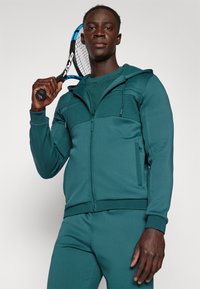Veste de sport zippée en bleu canard avec capuche, panneau texturé sur la poitrine et pantalon assorti. Modèle tenant une raquette de tennis, mettant en valeur le design sportif.