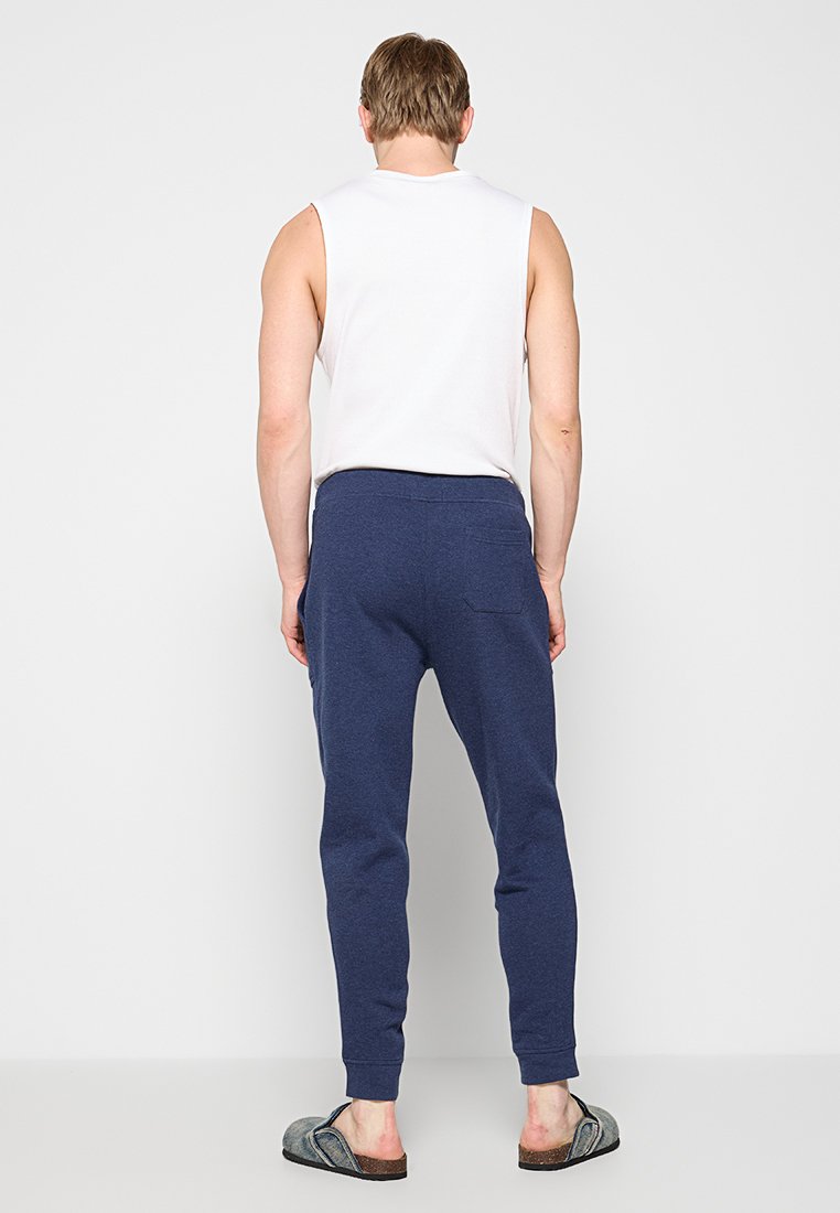 Granatowe dresy o luźnym kroju, miękkiej strukturze i elastycznym pasie, zestawione z białą koszulką bez rękawów oraz szarymi kapciami.