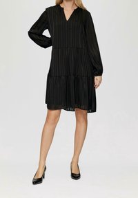 Černé šaty po kolena s vertikálními proužky, výstřihem do V a dlouhými rukávy. Mají vrstvený design a průsvitnou strukturu, nošené s černými lodičkami.