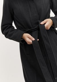 Cappotto di lana nera con una finitura tessurizzata, dotato di maniche lunghe e una cintura in tessuto. Chi indossa sta legando la cintura intorno alla vita.