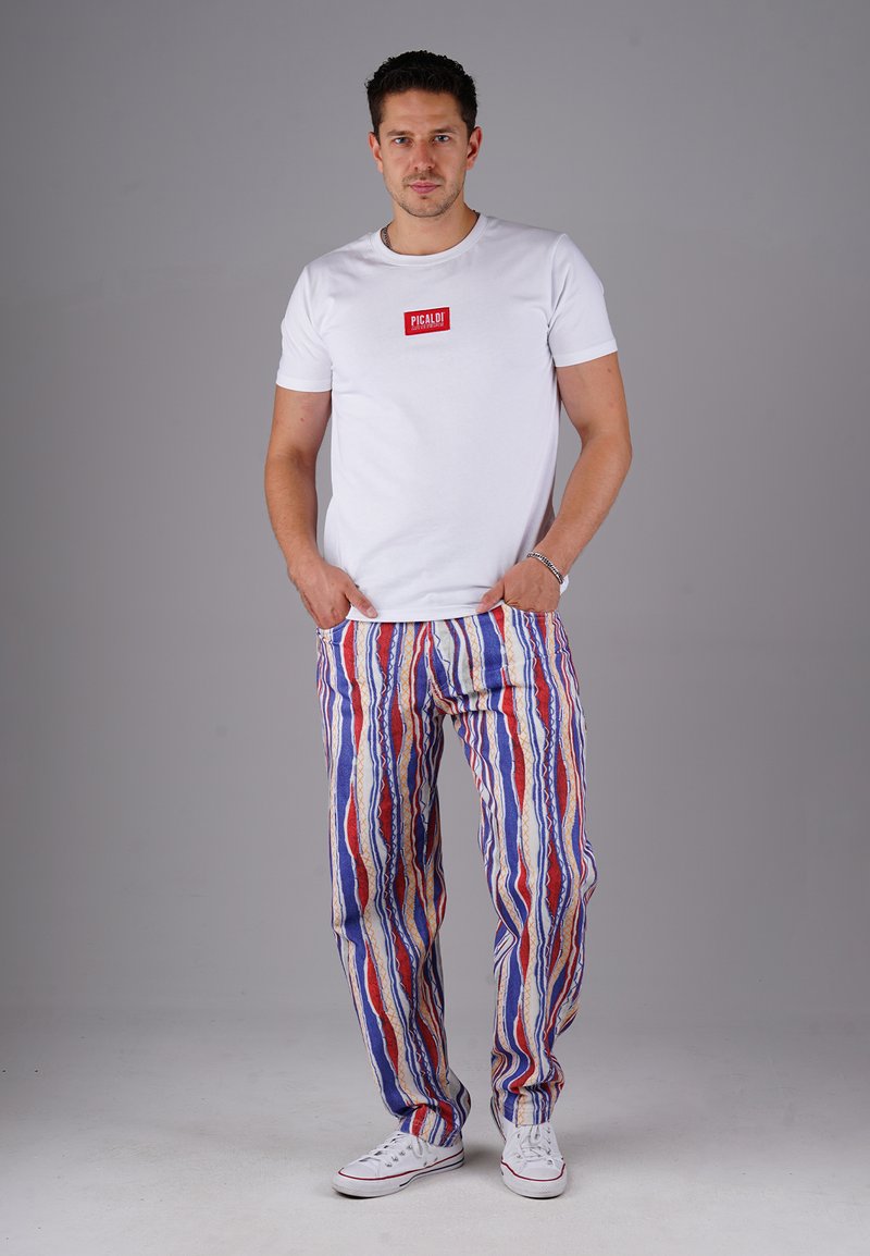 Weißes T-Shirt mit kurzen Ärmeln und rotem Logo, kombiniert mit bunten gestreiften Hosen in Blau-, Rot- und Goldtönen. Weiße Canvas-Sneaker.