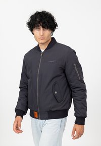 Donkerblauwe bomberjack met rits aan de voorkant, geribde manchetten en zoom. Voorzien van een voorvak en een bovenarmzak met klep.