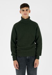 Maglione a collo alto verde scuro con texture a costine e polsini aderenti. Indossato con jeans grigi chiari, che mettono in risalto la robustezza del maglione.