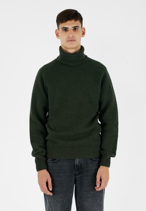 Maglione a collo alto verde scuro con texture a costine e polsini aderenti. Indossato con jeans grigi chiari, che mettono in risalto la robustezza del maglione.