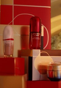Produkty pielęgnacyjne Shiseido w czerwono-złotym opakowaniu: przejrzysta szklana butelka z różową nakrętką, czerwona butelka oraz okrągły słoik z ombre.