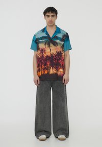 Camicetta a maniche corte con bottoni, di colore blu, con una stampa di un vibrante tramonto e palme, abbinata a pantaloni larghi in denim grigio.