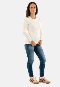 Maglione bianco lavorato a maglia con motivo testurizzato, jeans slim-fit blu e sneakers beige con strisce bordeaux. Il modello è in piedi con le mani sui fianchi.