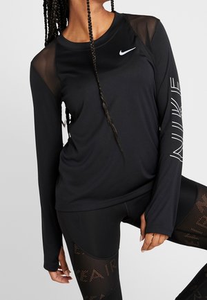 Osoba ubrana w czarną, długorękawową bluzę Nike z siateczkowymi ramionami i logo na rękawie, w połączeniu z czarnymi legginsami z siateczkowymi panelami i napisem "Air".