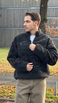 Μαύρο πουλόβερ από φλις με ψηλό γιακά, κλείσιμο με φερμουάρ και υφή με καφέ λεπτομέρειες. Φοριέται με ανοιχτό γκρι παντελόνι.