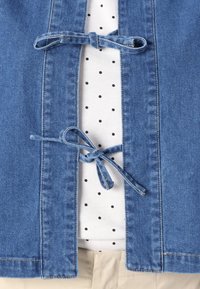 Veste en denim bleu clair avec fermeture à nouer, révélant une chemise blanche à pois noirs en-dessous et un pantalon beige.