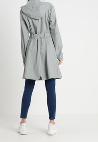 Jasnoszary, wodoodporny płaszcz przeciwdeszczowy z kapturem, taliowany, z rozcięciem z tyłu, w połączeniu z ciemnoniebieskimi obcisłymi dżinsami i białymi tenisówkami.