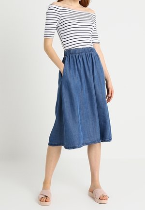 Femme portant un haut blanc à rayures noires et épaules dénudées, une jupe midi bleue taille élastique avec poches, et des sandales légères rose clair à enfiler.