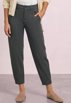 Pantalon ajusté gris foncé pour femme avec coutures avant et détail boucle en métal, assorti à des chaussures beiges à enfiler et une veste crème.