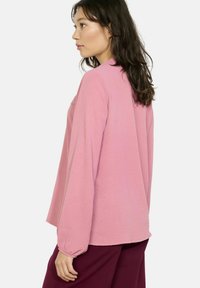 Różowa koszula z długim rękawem o luźnym kroju, z marszczonymi mankietami i subtelnym teksturowym wykończeniem z tyłu. Miękka tkanina, zaokrąglony dół.