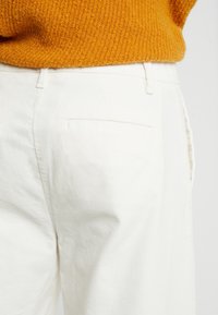 Vita bomullbyxor med rak benmodell, sidofickor och en siktad fåll. Komplementeras av en strukturerad senapsgul tröja.