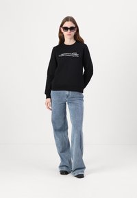 Sort sweatshirt med hvid tekst, paired med lyseblå flare jeans. Modellen bærer sorte solbriller og har langt brunt hår.