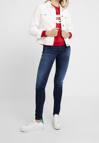 Chaqueta de mezclilla blanca sobre un sudadera roja con texto blanco, combinada con jeans ajustados azul oscuro y zapatillas blancas con acentos en azul teal.