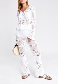 Ensemble deux pièces en maille blanche comprenant un haut ample à manches longues avec un tissage ouvert et un pantalon évasé avec un motif similaire. Sandales et sac rond inclus.