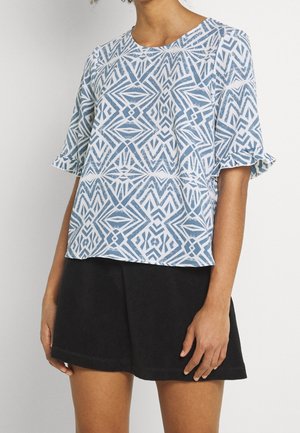 Kvinde iført en kortærmet bluse med blå og hvide geometriske mønstre og flæset ærmer samt et sort skørt.