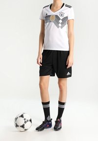 Bílý fotbalový dres s černými pruhy a zlatým emblémem, doplněný černými šortkami a pruhovanými černými ponožkami. Fotbalový míč na zemi.