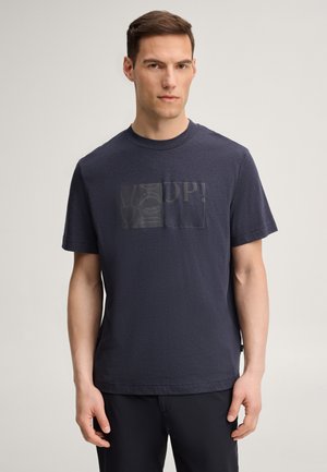 Homme debout devant un fond uni, portant un T-shirt bleu marine à manches courtes avec un motif géométrique tonal et un texte sur la poitrine.
