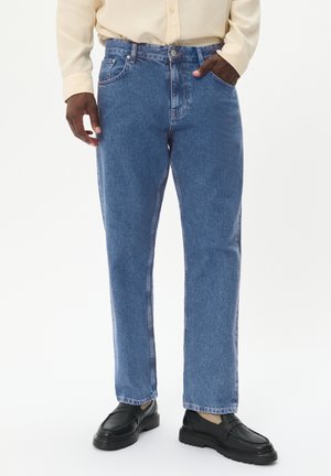 Jeans de mezclilla azul con un corte relajado, que cuentan con una cremallera, diseño de cinco bolsillos y un leve desvanecimiento. Llevados con zapatos negros tipo slip-on.