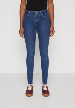 Kobieta ubrana w niebieskie obcisłe dżinsy, czarny bezrękawnik, białe skarpetki i bordowe trampki z białymi sznurowadłami stoi na jednolitym tle.