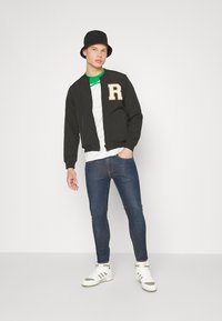 Casaco bomber preto com a letra "R" em tom de areia, t-shirt branca, jeans skinny azul-escuro e ténis brancos com detalhes verdes, usados com um chapéu bucket preto.