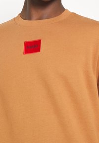 Kurzärmliger Sweatshirt mit Rundhalsausschnitt aus hellbraunem Stoff. Verfügt über ein rotes rechteckiges Patch mit "HUGO" in schwarzer Schrift. Glatte Textur.
