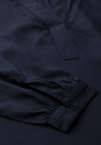 Blusa azul marino con escote en V y mangas largas con puños fruncidos. Fabricada con tejido suave, incluye un botón en el puño.