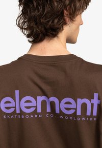 Camiseta de algodón marrón con texto grande en púrpura que dice "element" y "SKATEBOARD CO. WORLDWIDE" en la parte de atrás. Diseño de manga corta.