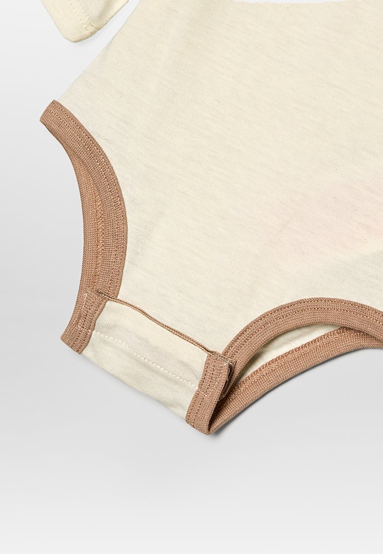 Mono de color crema con mangas cortas, con ribetes marrones. Material de algodón suave con una ligera textura, diseño ajustado y cierres a presión.