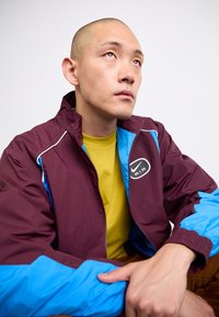 Man med en vinröd och blå Nike Air-jacka och gul skjorta, sittande och tittande uppåt mot en vit bakgrund.