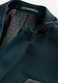 Mörk teal jacka med strukturtyg, med ett mönstrat svart foder. Kragen har en mjuk detalj som visar på hantverkets kvalitet.